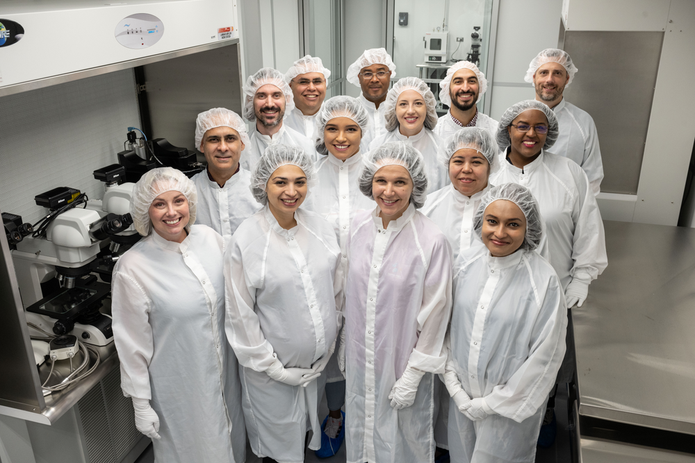 Fourteen people dressed in white gowns pose for a photo inside a laboratory.